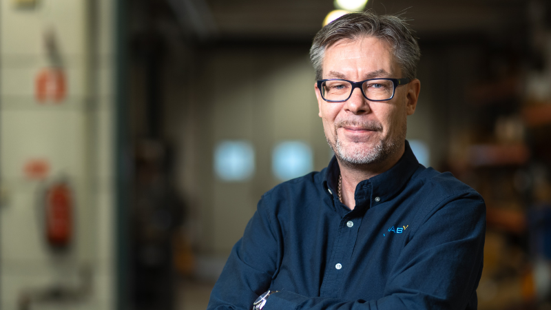 VIMAB-medarbetare med korsade armar i verkstadsmiljö, iförd mörkblå arbetsskjorta med VIMAB-logotyp
