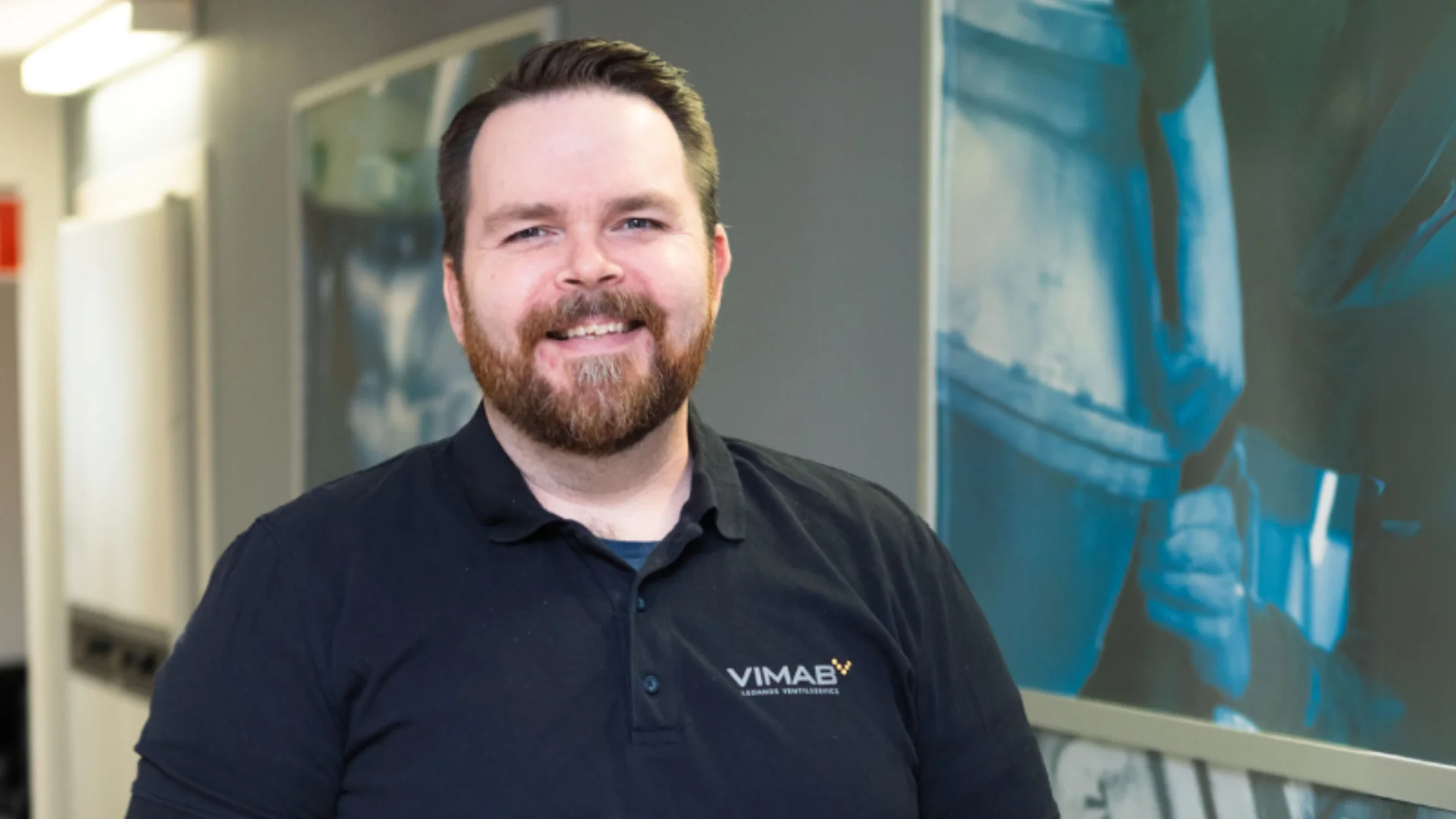 VIMAB-medarbetare ler mot kameran i kontorsmiljö med VIMAB-logotyp på arbetsskjortan