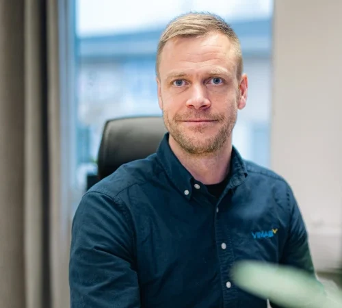 Möt Patrik: En man med kort blont hår och skägg sitter vid ett skrivbord på ett kontor, klädd i en mörkblå skjorta med knappar och tittar in i kameran, med ett fönster och gardiner i bakgrunden.