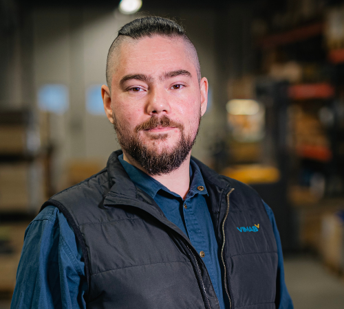 Möt Dan, en person med skägg och mustasch, klädd i blå skjorta och svart väst, står i en lagerlokal fylld med hyllor och lådor. Bakgrunden är något suddig.